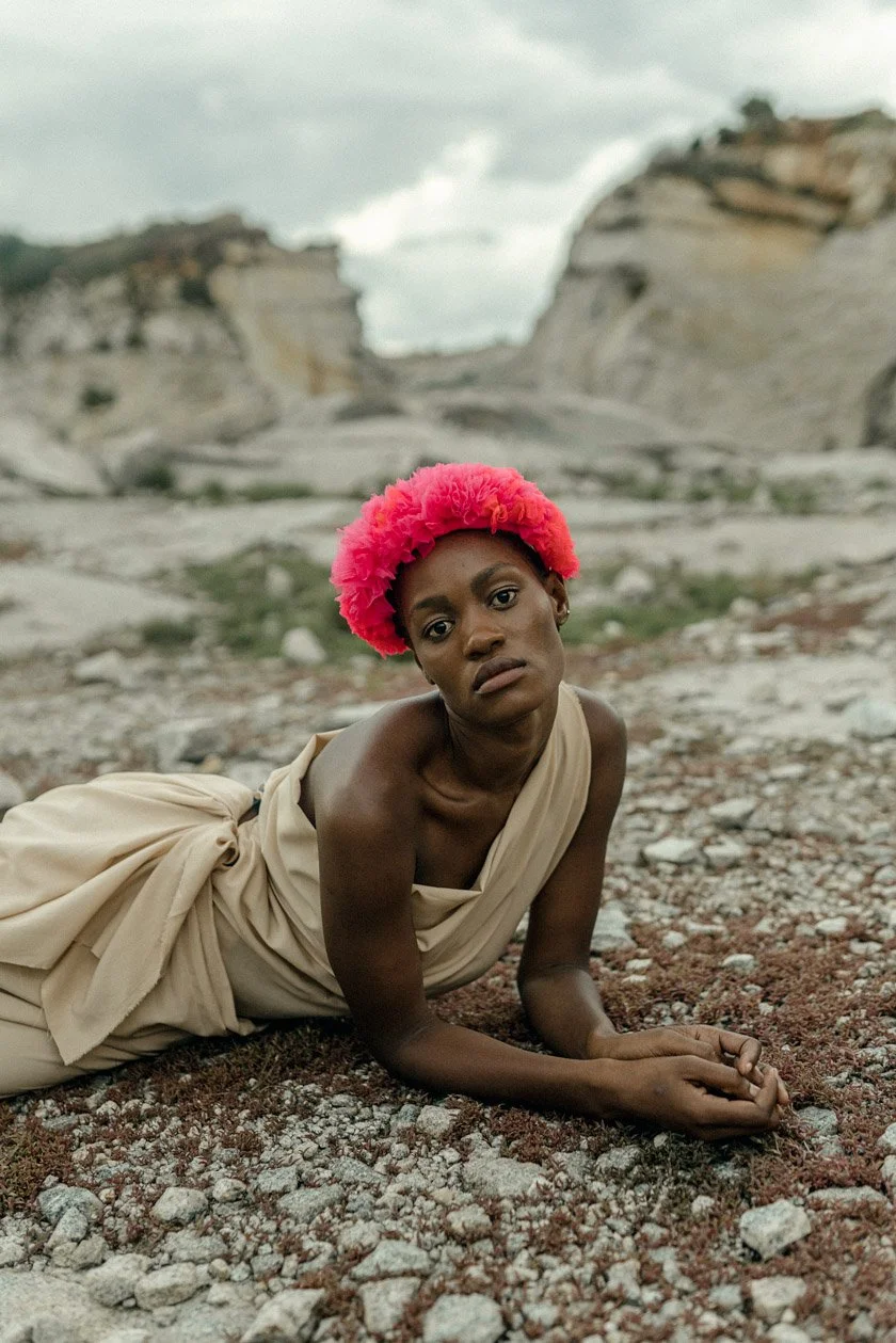 A woman with dark skin and a vibrant pink afro hairstyle, wearing a beige draped garment, lying on rocky terrain in a mountainous landscape under cloudy skies.