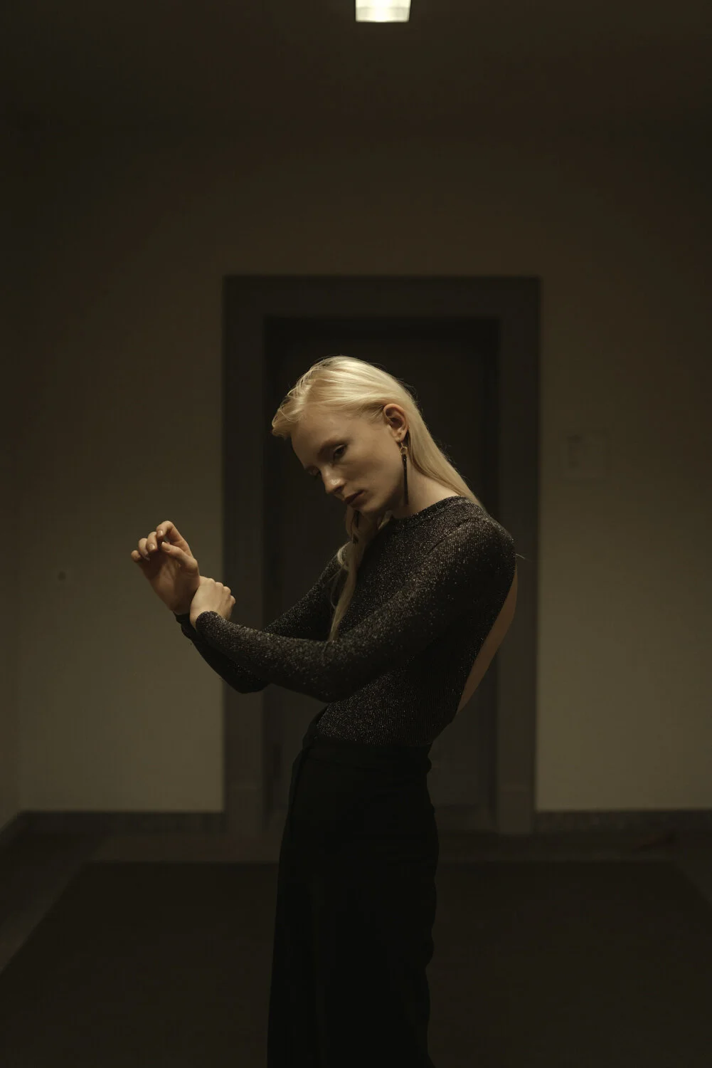 A woman with long blonde hair posing indoors, wearing a black, sparkly, long-sleeved top and black high-waisted pants, striking a contemplative pose.