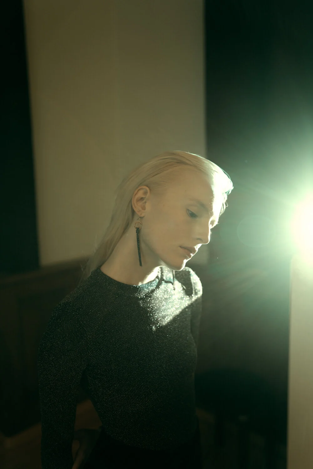 A woman with platinum blonde hair wearing a black, sparkly top and long black earrings, illuminated by bright light from the right side.