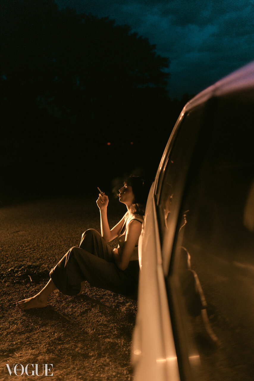 A woman sitting on the ground at night beside a car, smoking a cigarette, with her reflection visible on the car window, under a dark, cloudy sky.