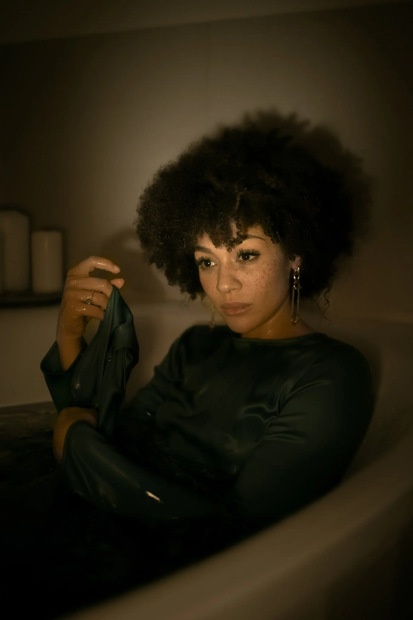 A woman with curly hair, freckles, and hoop earrings, sitting in a dimly lit room, holding a black leather jacket.