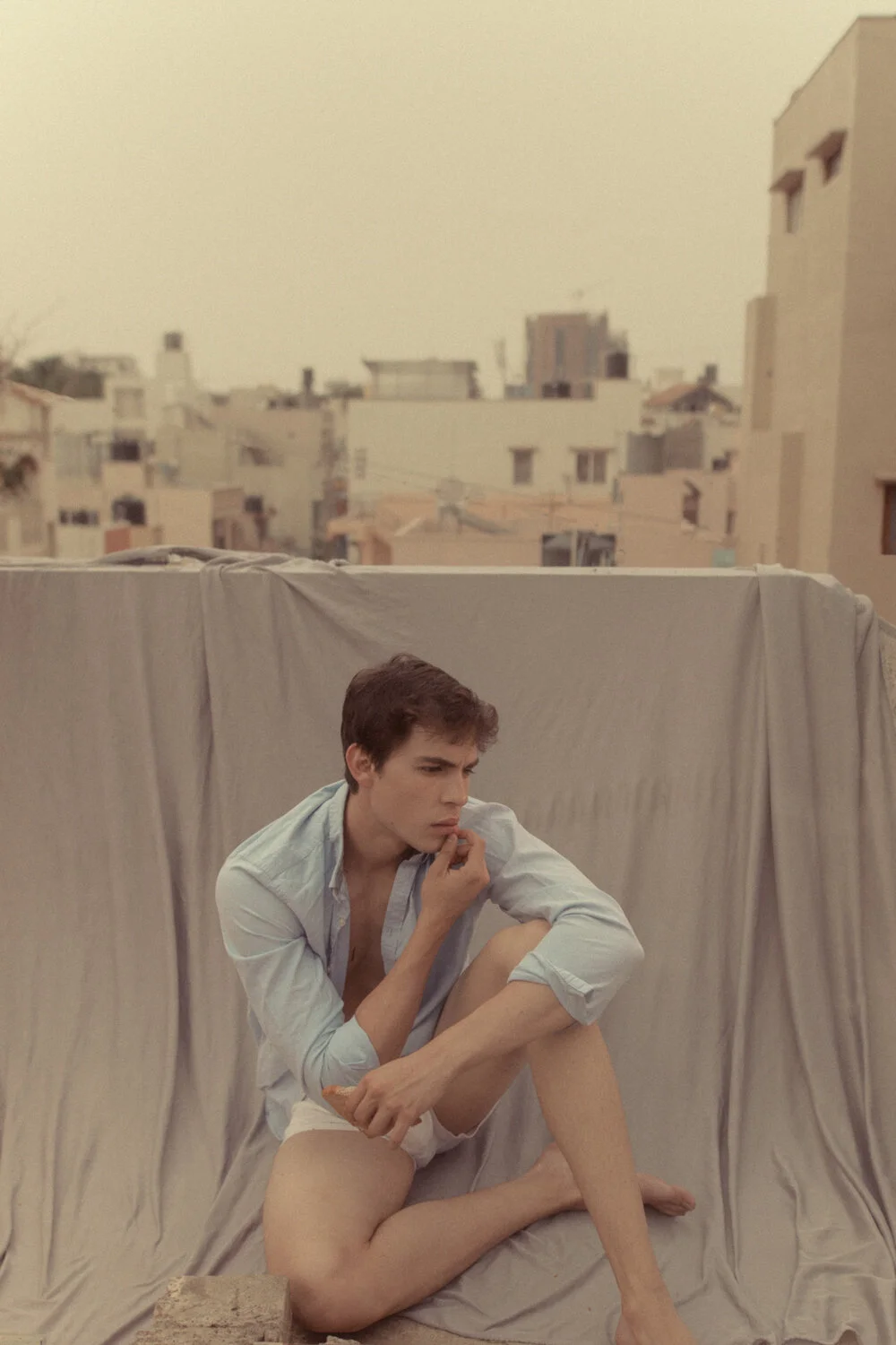 A young man sitting cross-legged on a makeshift sofa made from a sheet on a rooftop, with an urban cityscape in the background, wearing a button-up shirt and shorts, in a pensive pose.