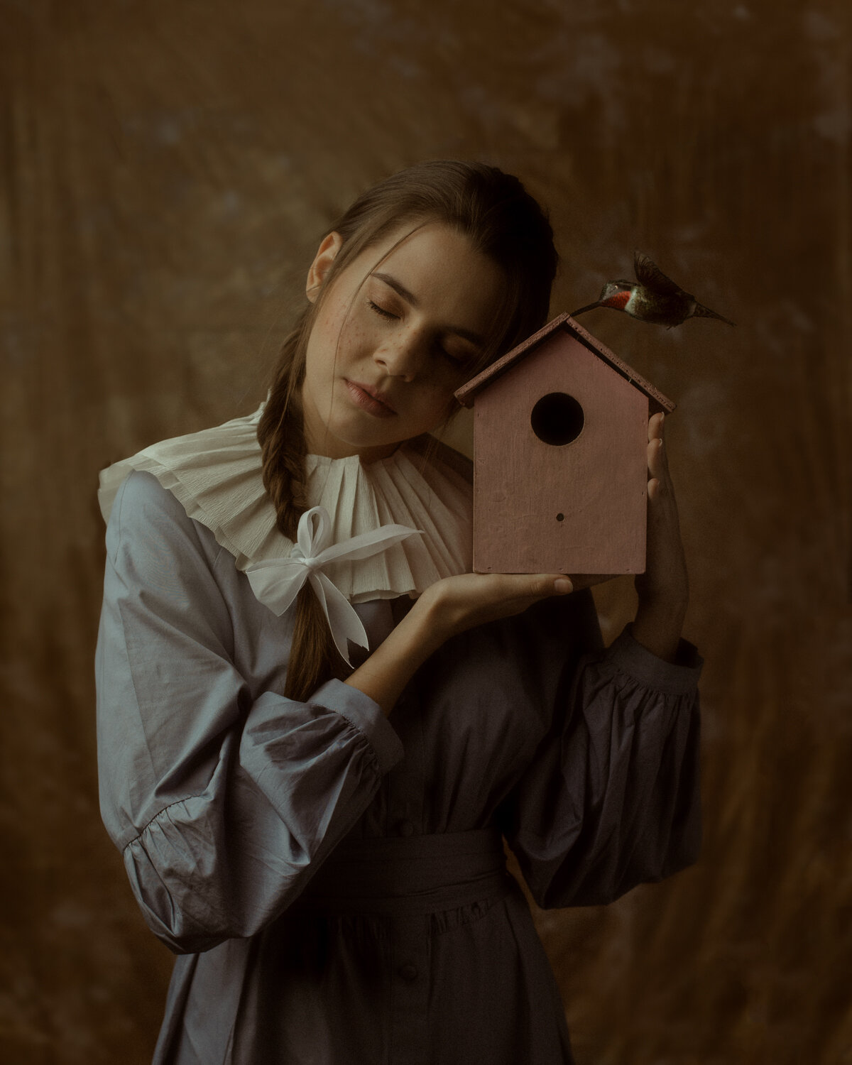 A woman with long hair in a braid is holding a pink birdhouse with a bird on top, her eyes closed as if she is cradling it gently.