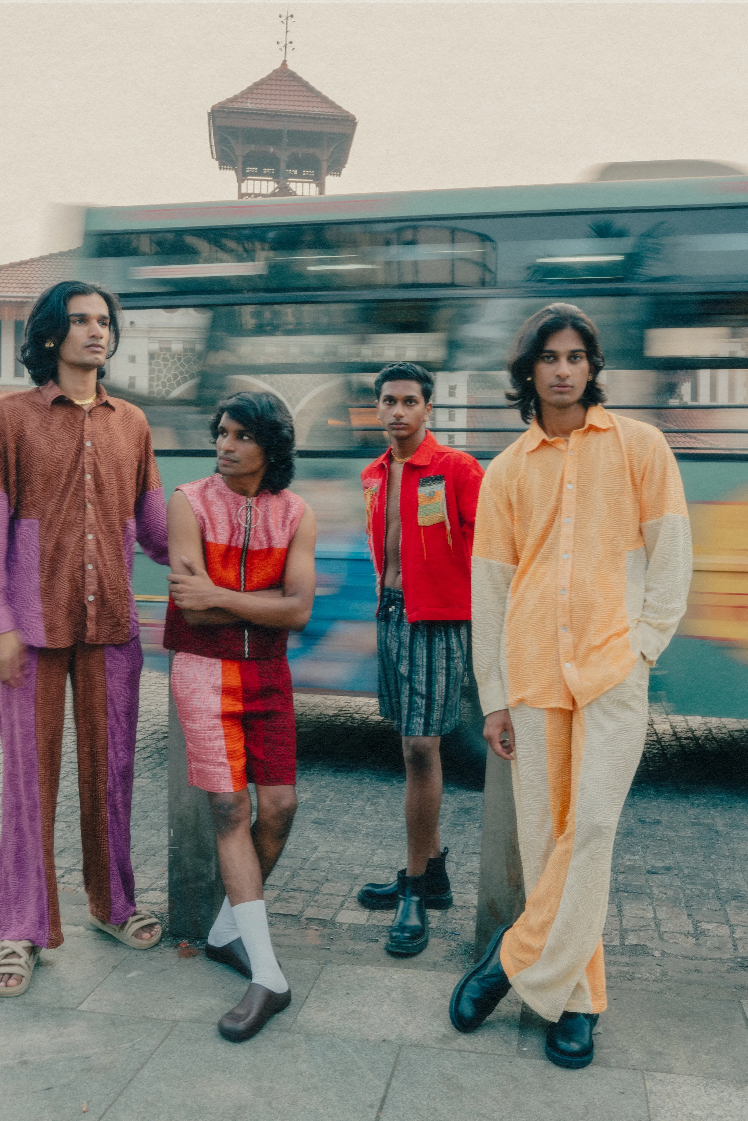 Four young men standing in front of a blurred moving bus on a city street, with historic buildings in the background.