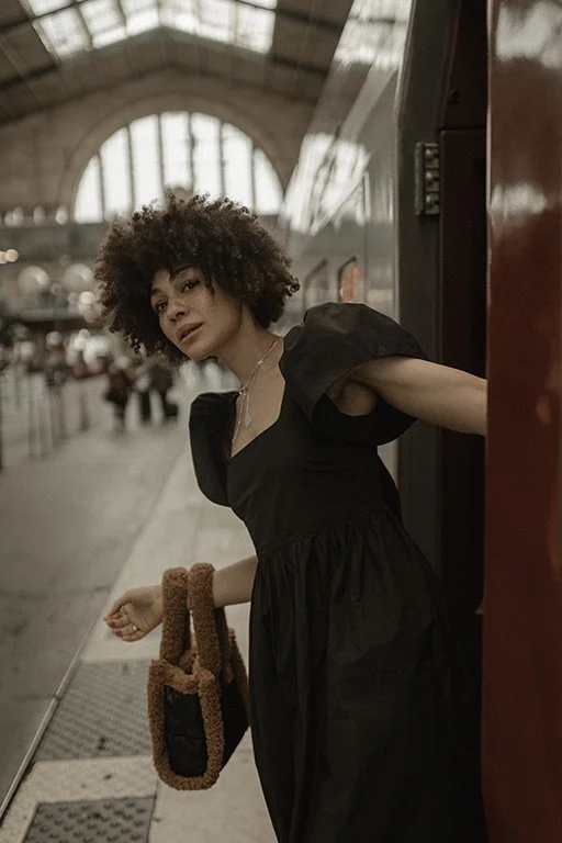 Woman with curly hair in a black dress leaning out of a train door at a train station.