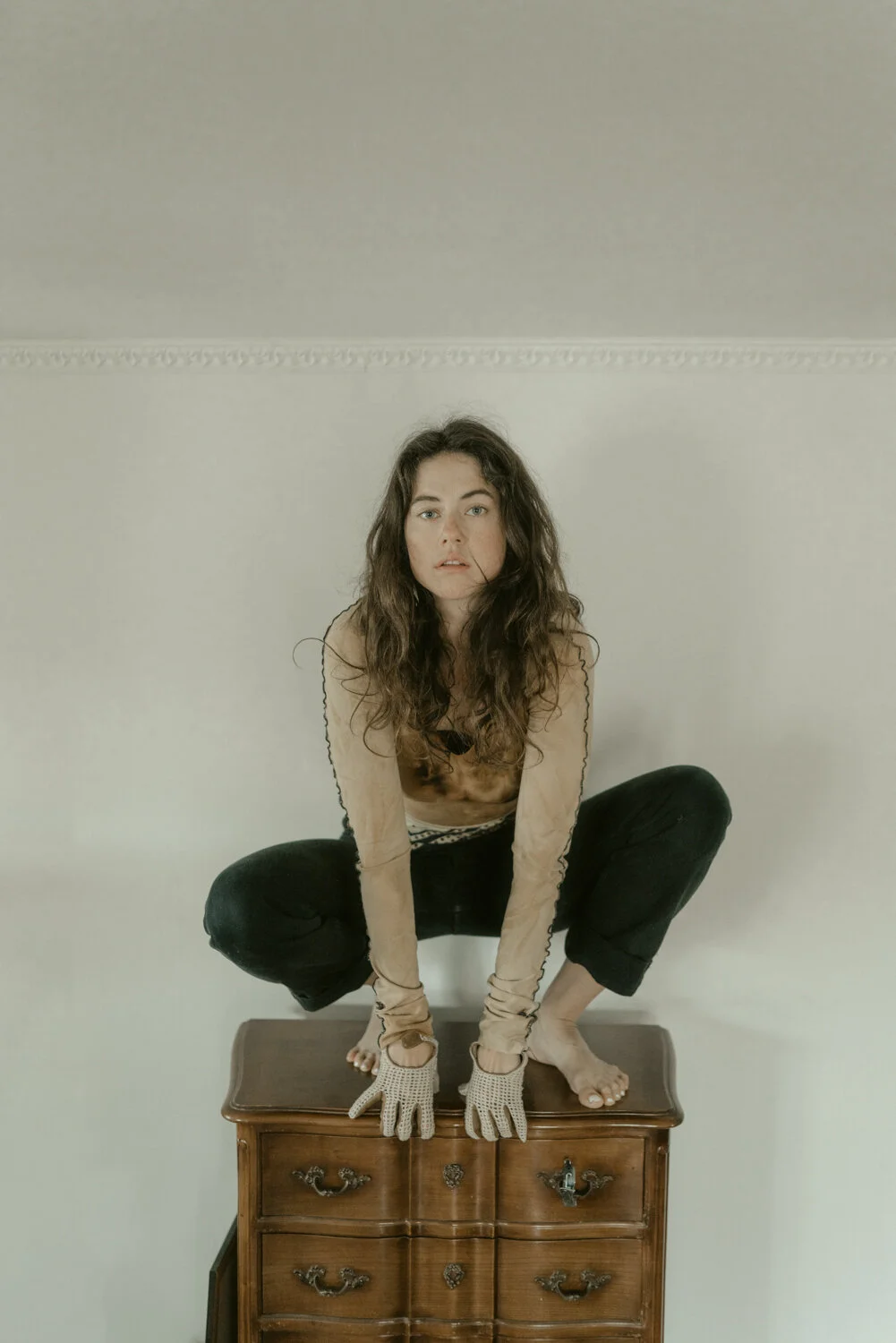 A woman with long, wavy hair crouching on a small wooden dresser, wearing gloves, beige top, and black pants with a patterned waistband, in a minimalistic room with a plain wall and ceiling.