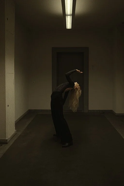 A person with long blonde hair wearing dark clothing bending backward in a dimly lit hallway with a fluorescent ceiling light.