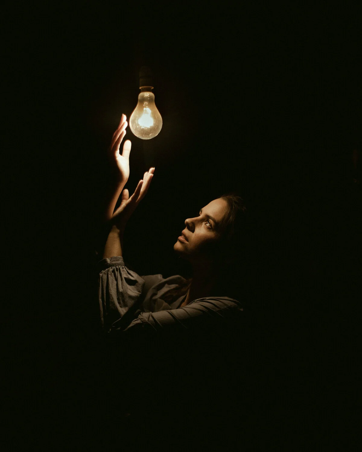 A woman reaches towards a glowing light bulb hanging from the ceiling in a dark room.