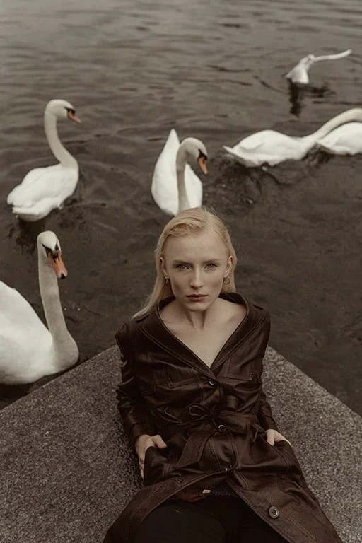 A woman with blonde hair sitting on a stone ledge near water with swans swimming behind her