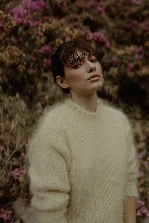 A woman with dark wet hair and wearing a cream-colored sweater stands outdoors in front of pink flowers and shrubbery.