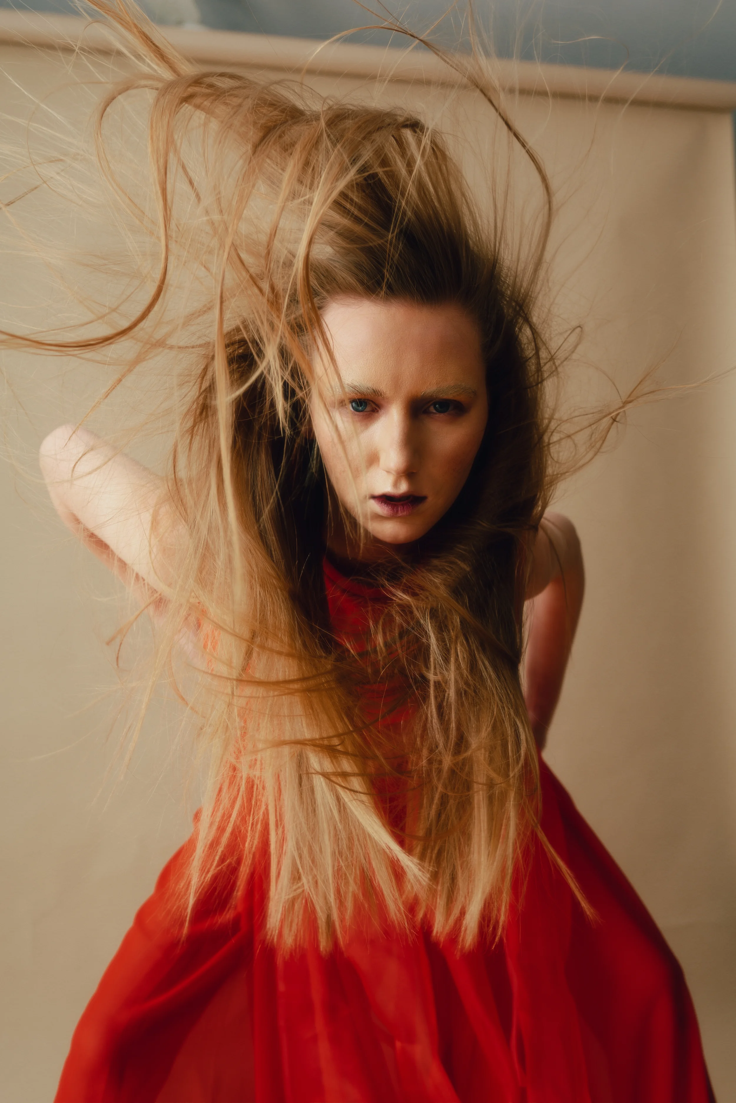 A woman with long, tousled red hair wearing a red dress, looking intensely at the camera in a close-up shot.