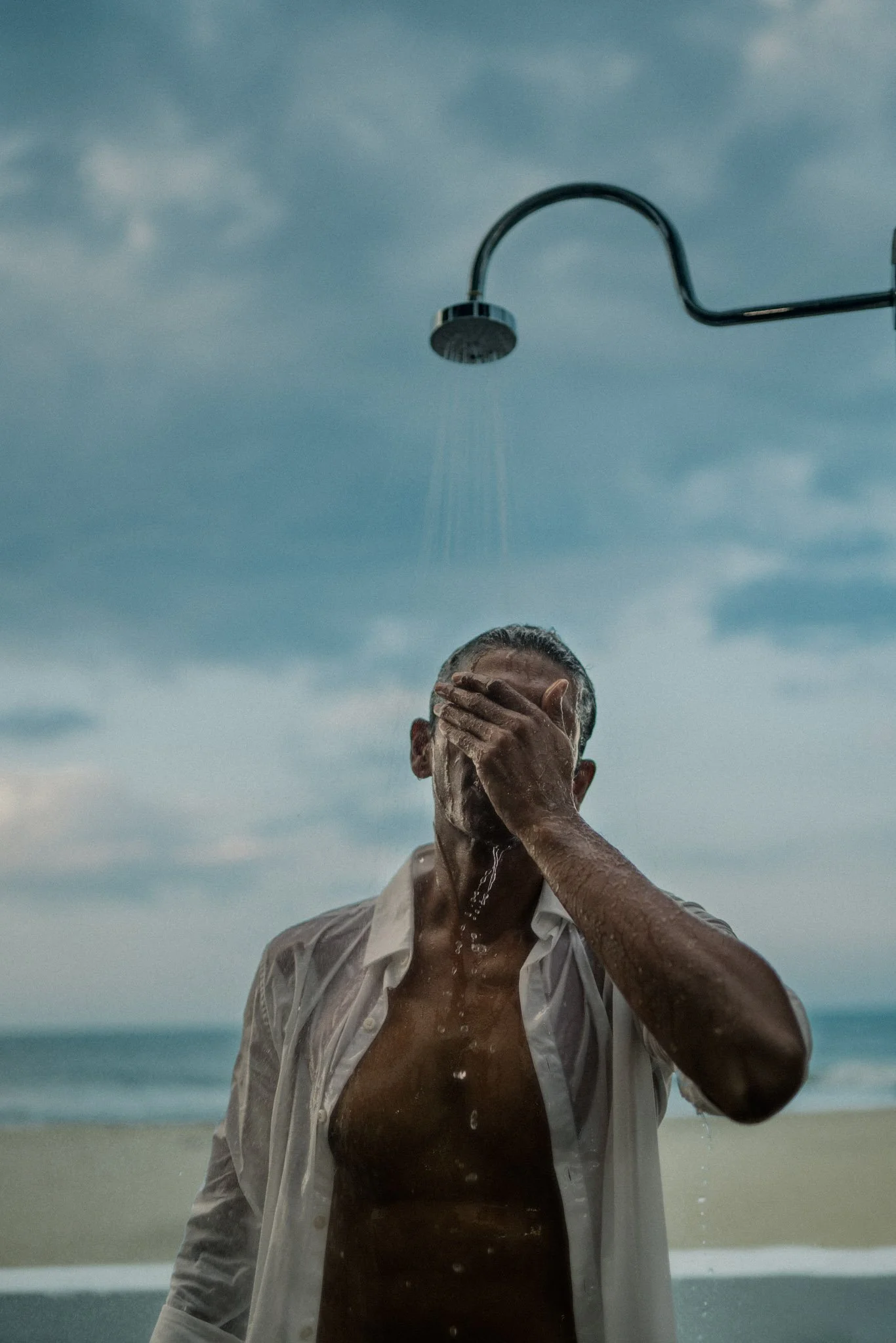 A man washing his face under an outdoor shower on the beach with a cloudy sky in the background.