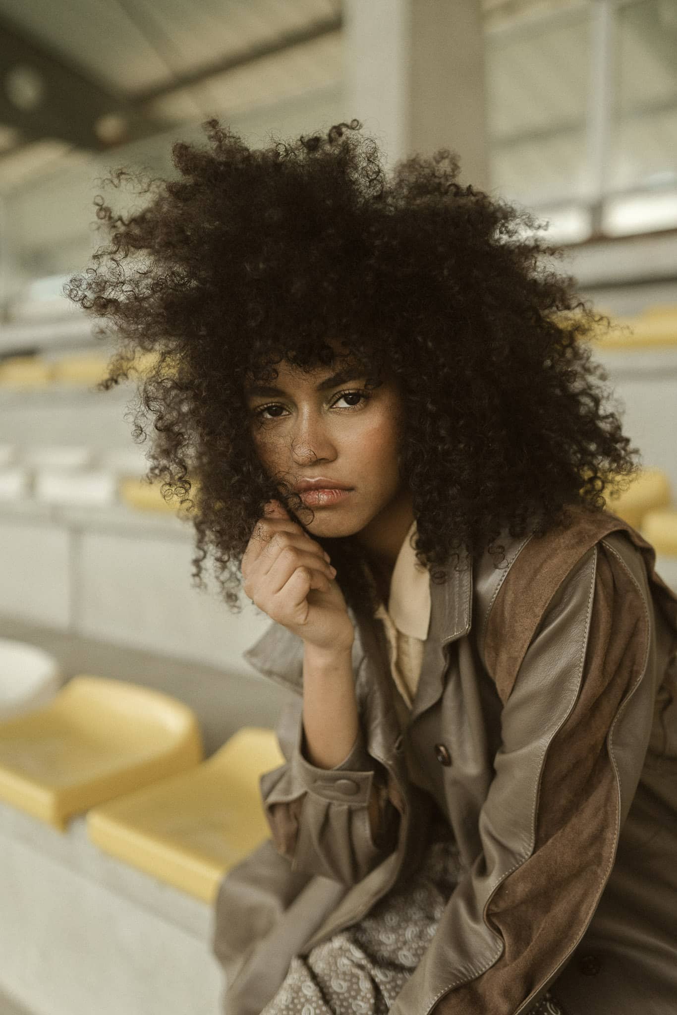 A young woman with curly black hair, sitting on a yellow stadium seat, in an indoor setting, wearing a brown jacket and a beige shirt, with her chin resting on her hand, looking at the camera.
