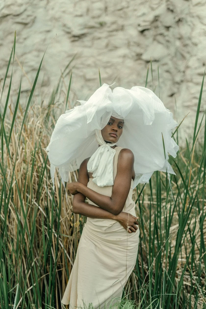 A woman standing among tall green reeds with a rocky background, wearing a beige dress and a large white fabric hat.