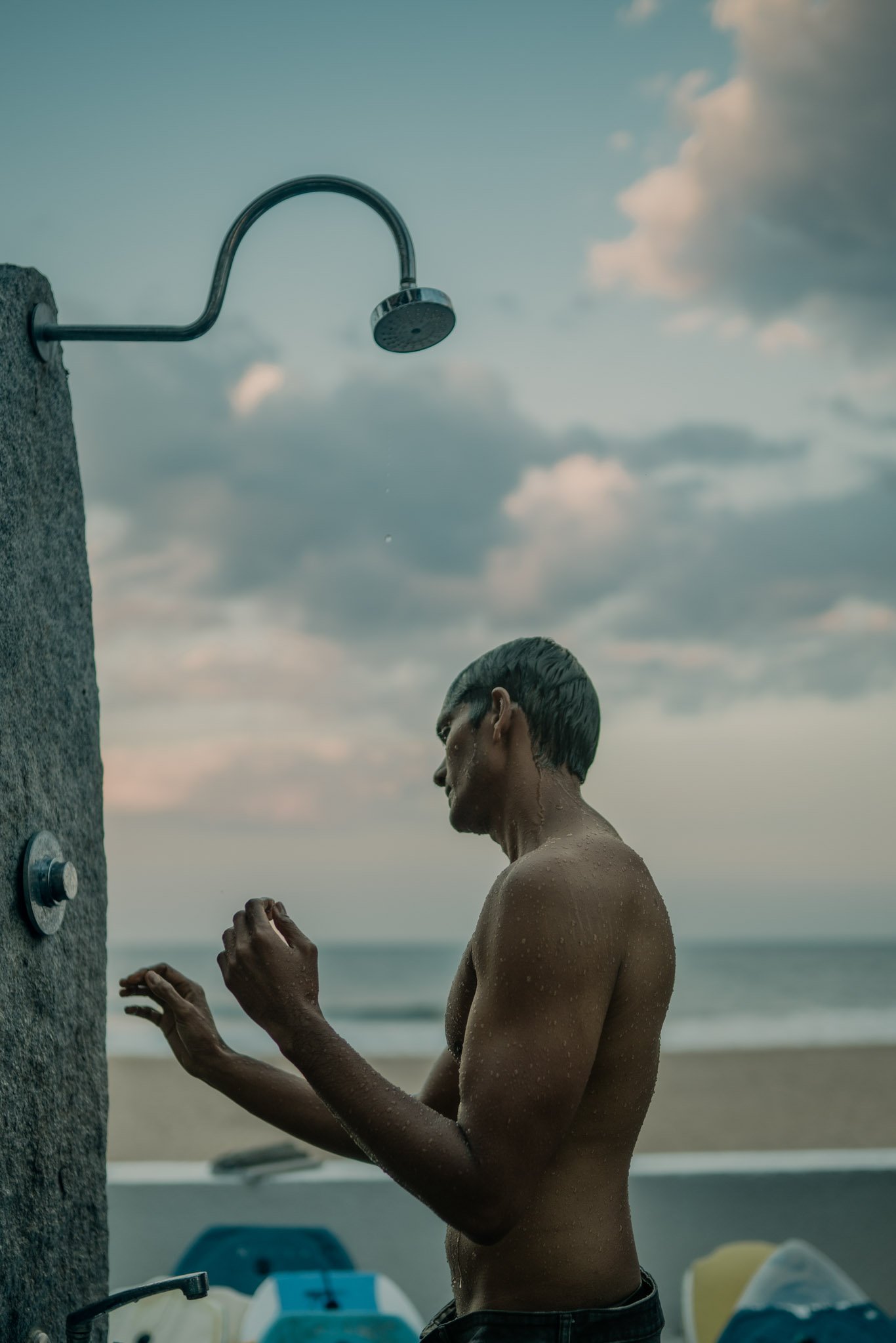 A man with wet hair is standing under an outdoor shower on a beach during sunset or sunrise, with a cloudy sky in the background.