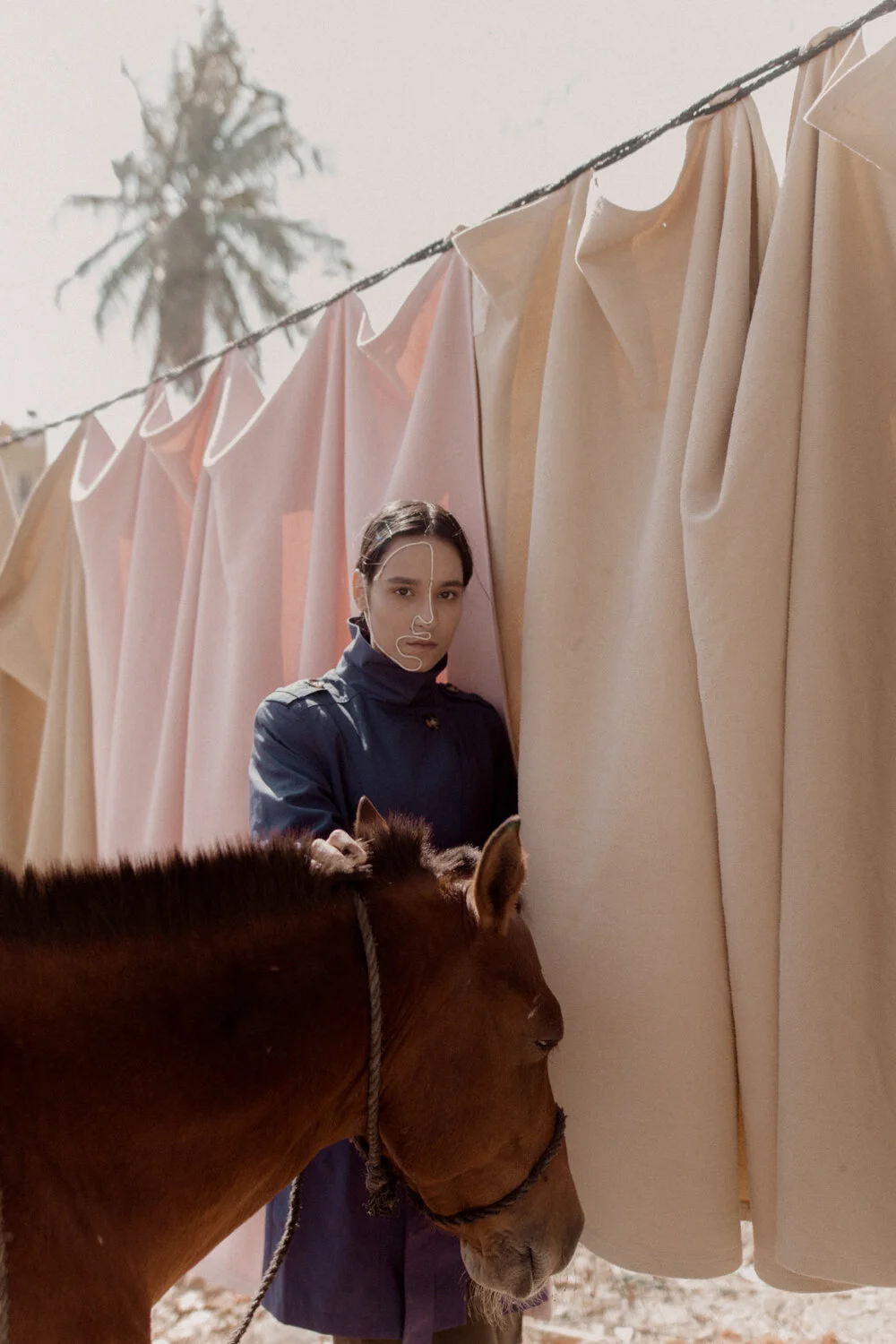 A woman with dark hair, dressed in a dark blue jacket, stands among beige and pink curtains hanging on a line with a blurred palm tree in the background. She has a minimalist white line drawing of a face on her face. A small brown horse with a rope h