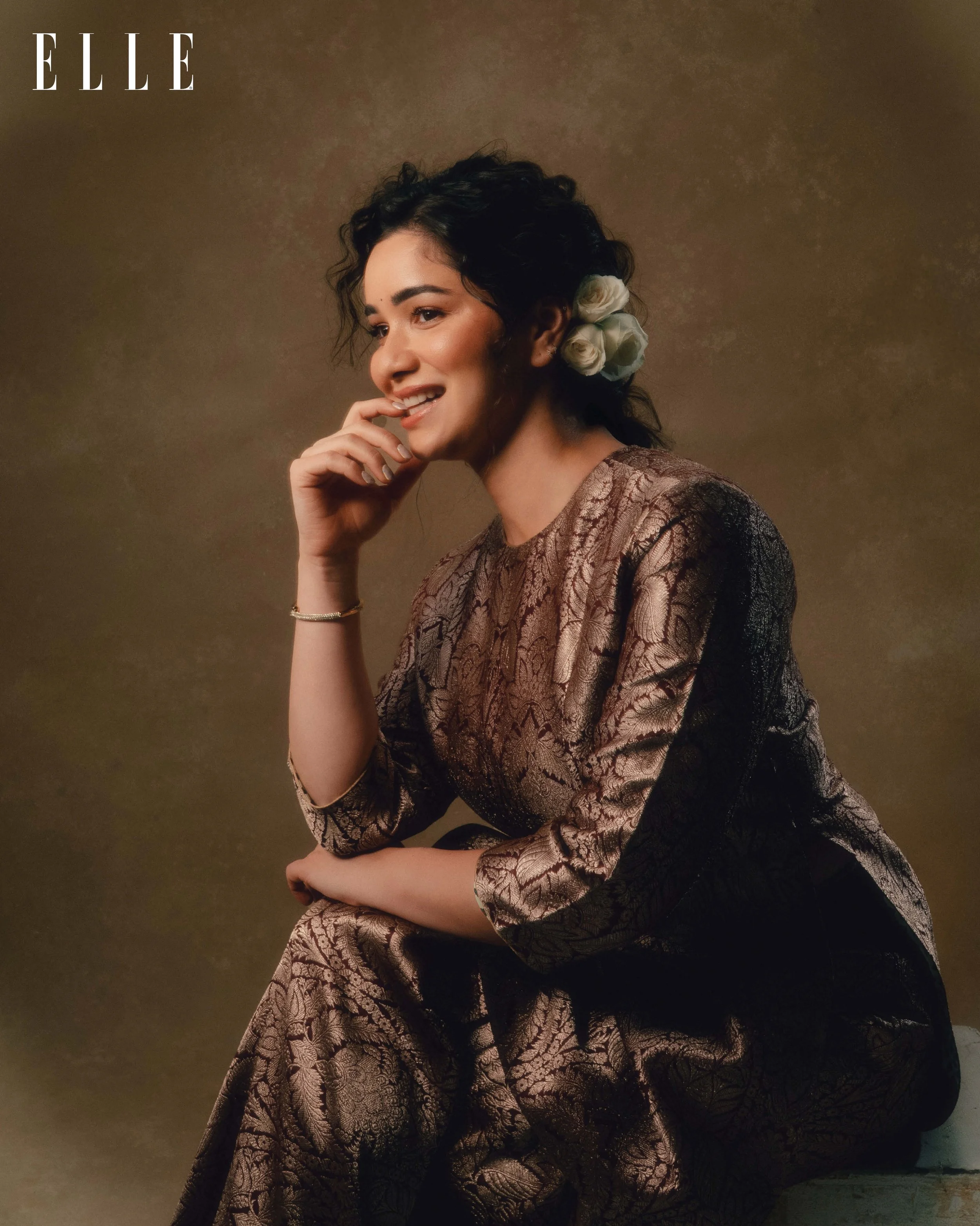 A woman with dark curly hair decorated with white roses, wearing a shiny patterned dress, sitting with her chin resting on her hand and smiling, against a warm brown background, with ELLE magazine logo in the top left corner.