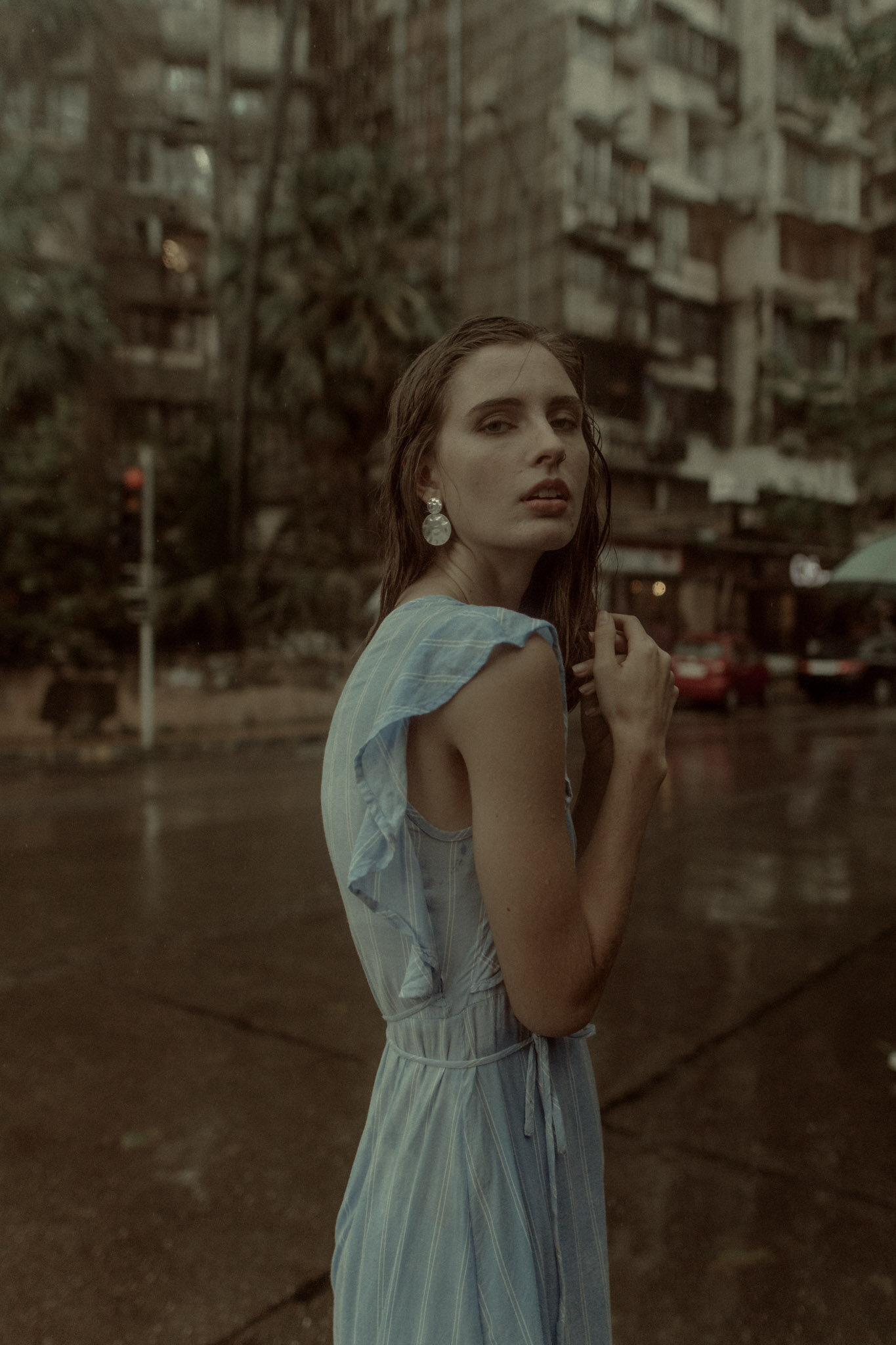 A young woman with wet hair and statement earrings stands on a city sidewalk, looking at the camera, with an apartment building and trees in the background on a rainy day.