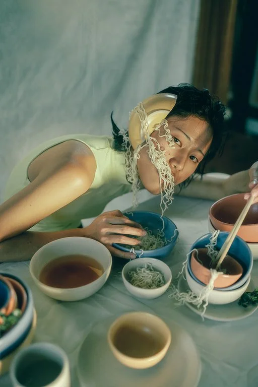 A young woman lying at a table with various bowls of ramen and tea, holding chopsticks with noodles hanging from her mouth.