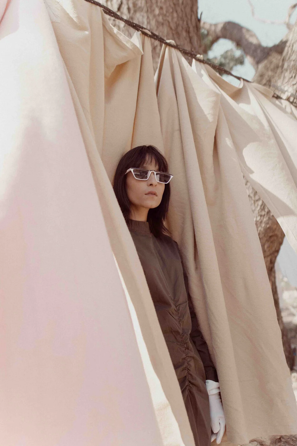 A woman with shoulder-length dark hair and sunglasses looks to the side while standing behind beige curtains hanging from a tree branch outdoors.