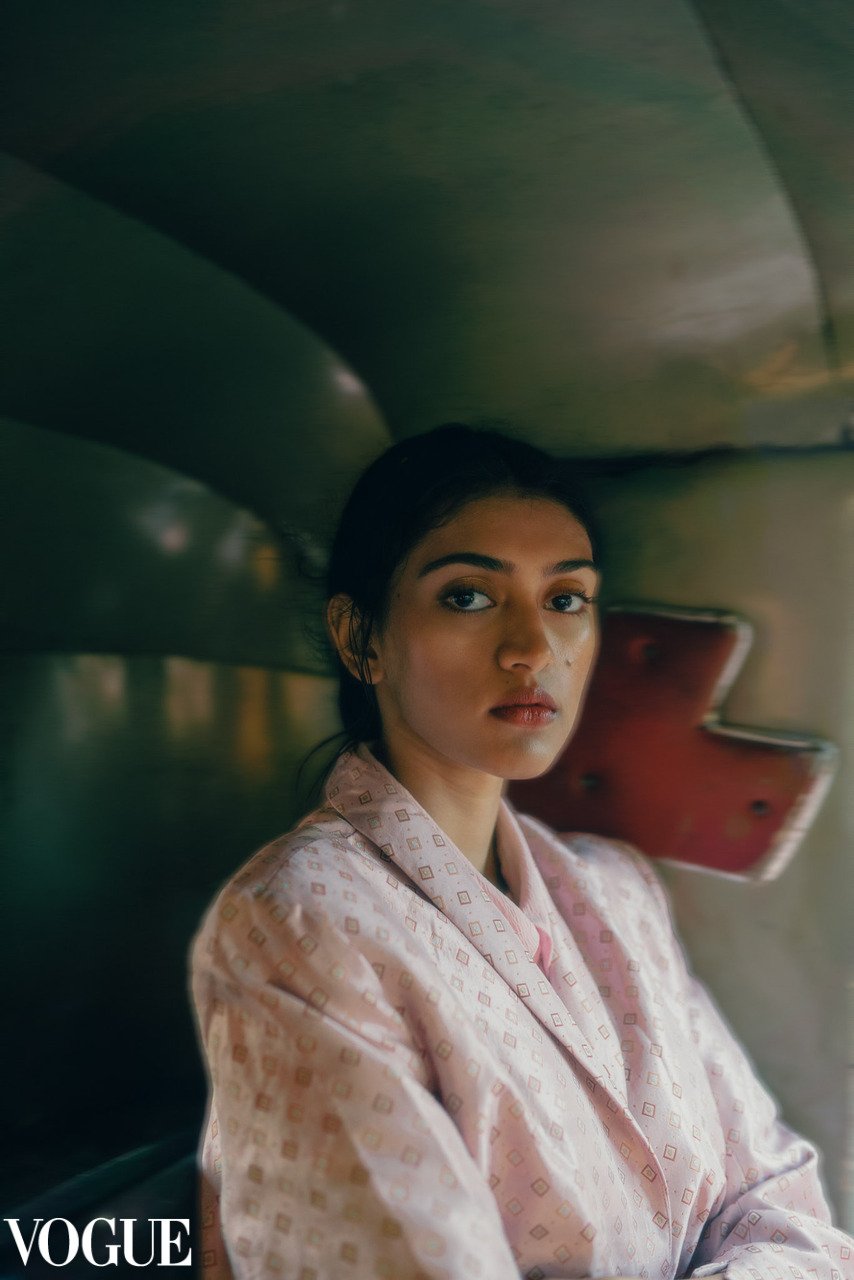 A woman with dark hair is sitting, wearing a light pink patterned silk robe. She is looking directly at the camera with a neutral expression, inside a room with a curved ceiling and a red piano in the background.