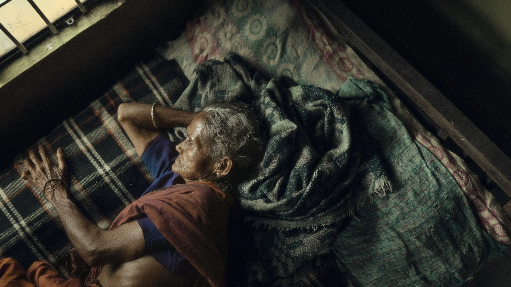 An elderly woman sleeping in a bed, lying on her side with her hand resting at her side. She is covered with a blanket and wearing a blue shirt.