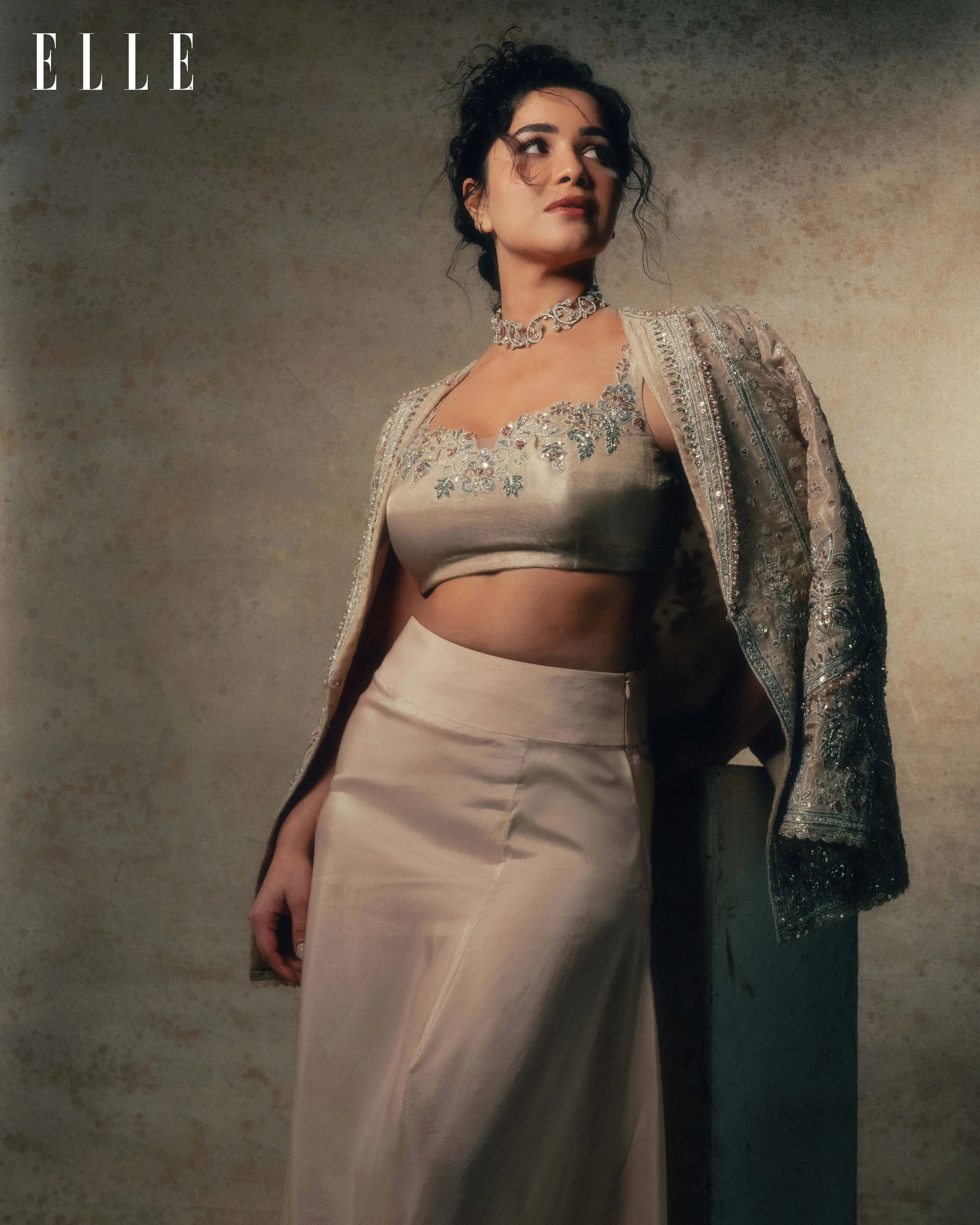 A woman wearing a beige embroidered blouse and skirt, with a matching embroidered cape, accessorized with a necklace, posing against a textured beige background.