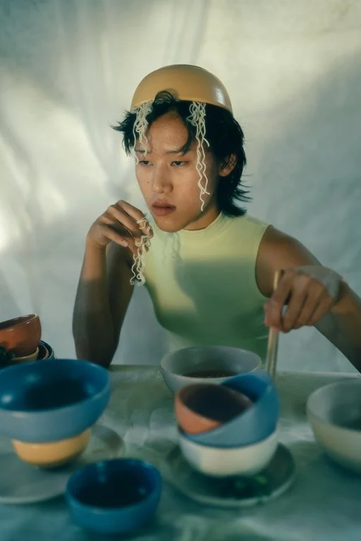 A young person with dark hair, wearing a yellow sleeveless shirt and a beige bowl on their head, is sitting at a table with colorful bowls and plates, eating noodles with chopsticks.