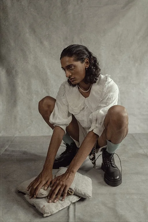 Person squatting on the floor, wearing a white blouse, black boots, gray socks, and a necklace, pressing down a cloth-covered object with their hands in a minimalist room with a gray wall and floor.