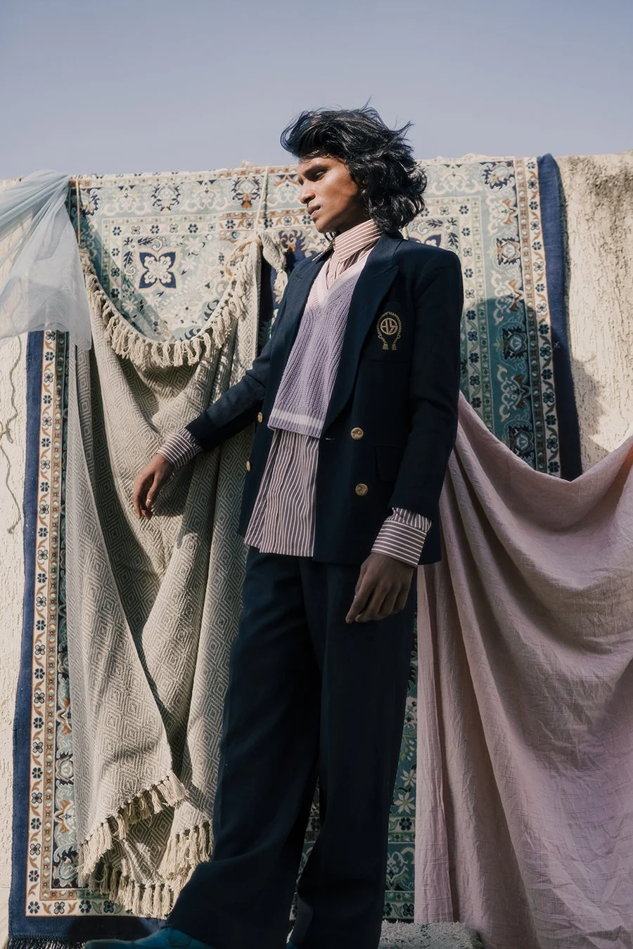 Person with dark, tousled hair wearing a navy blazer with a crest patch over a striped shirt, standing in front of patterned textiles outdoors under a clear sky.
