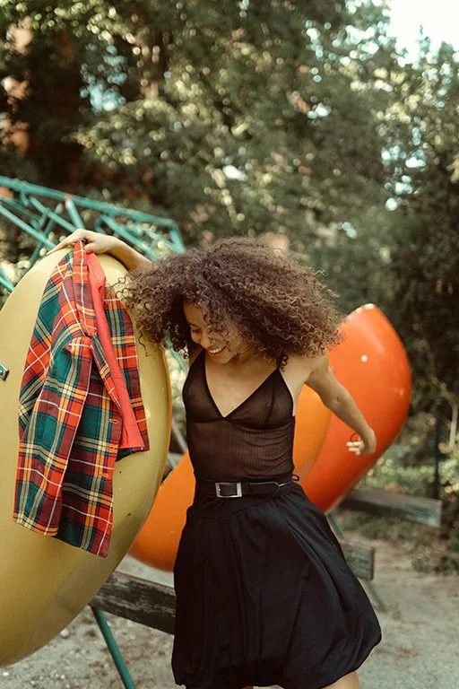 A woman with curly hair in a black dress smiling and leaning against colorful outdoor chairs in a park.