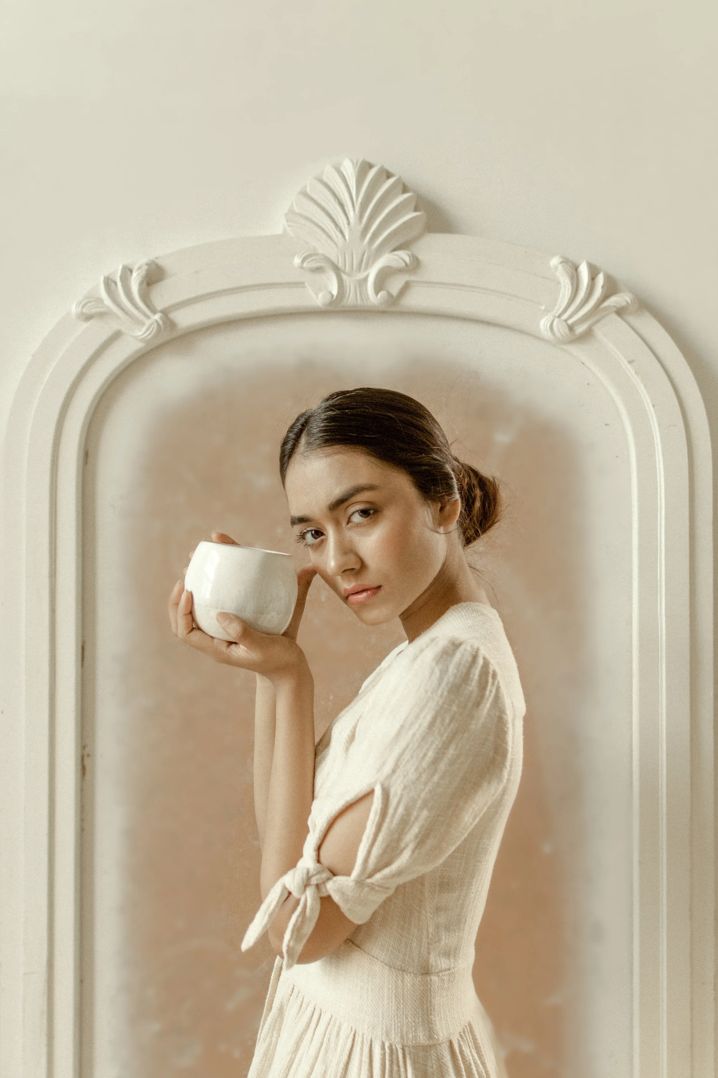 A woman with dark hair tied back in a low bun holding a cream-colored mug, standing in front of a decorative mirror with ornate white frame.