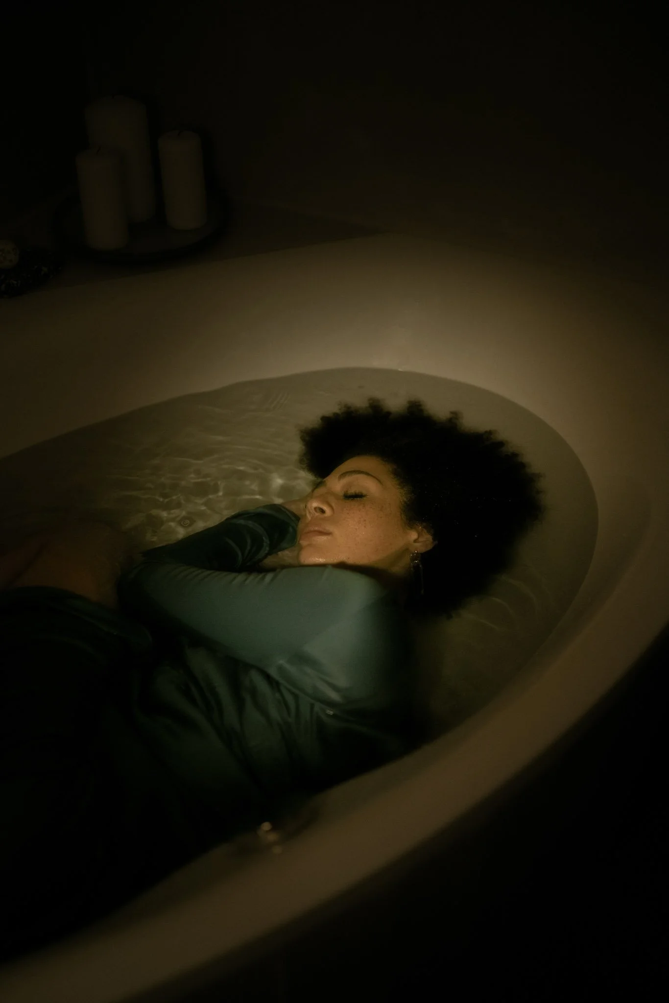 A woman with curly hair is lying in a bathtub filled with water, with her eyes closed, appearing relaxed.