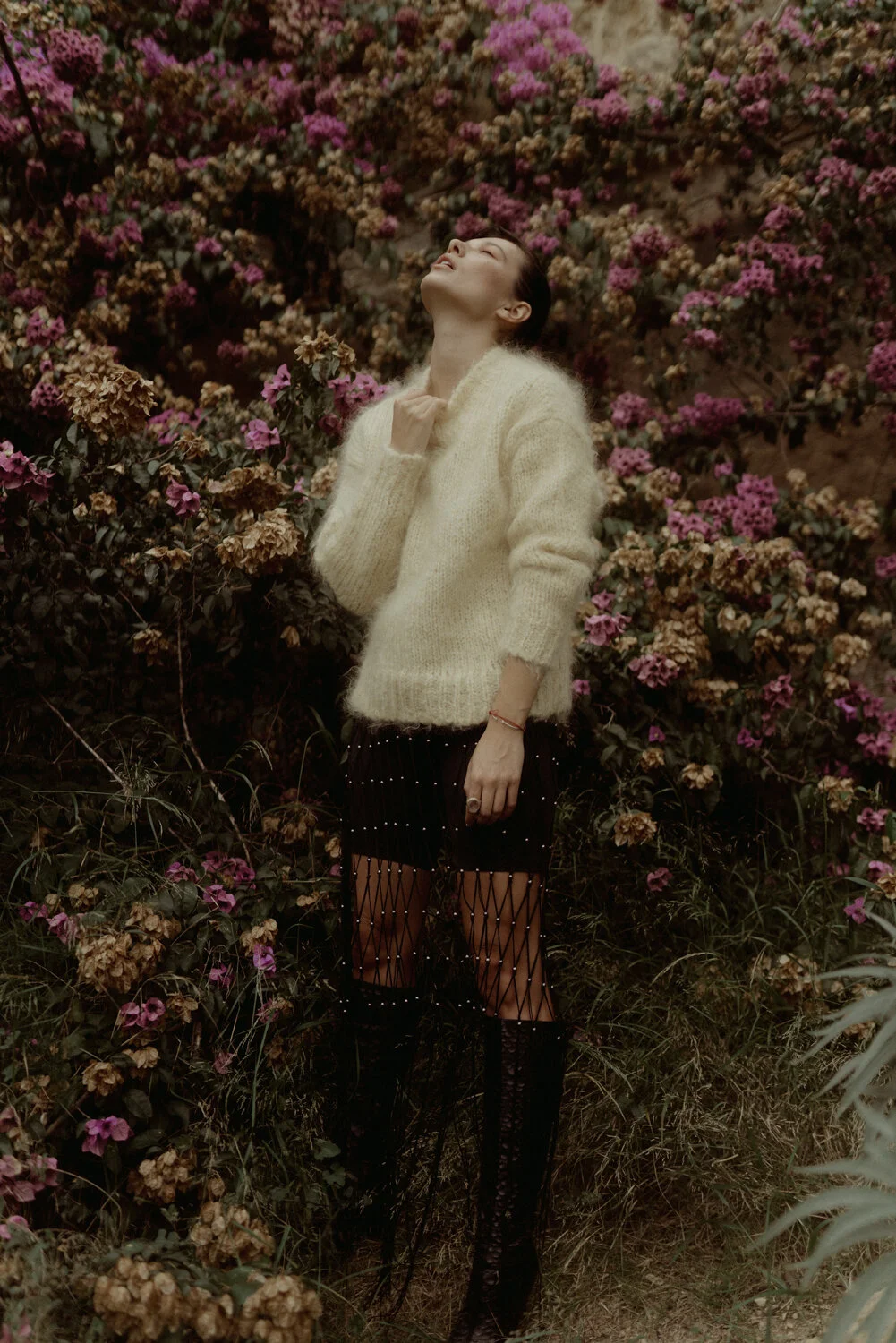 A woman in a cozy cream sweater and fashionable black fishnet pants stands amidst blooming pink and brown flowers with her eyes closed and head tilted up.