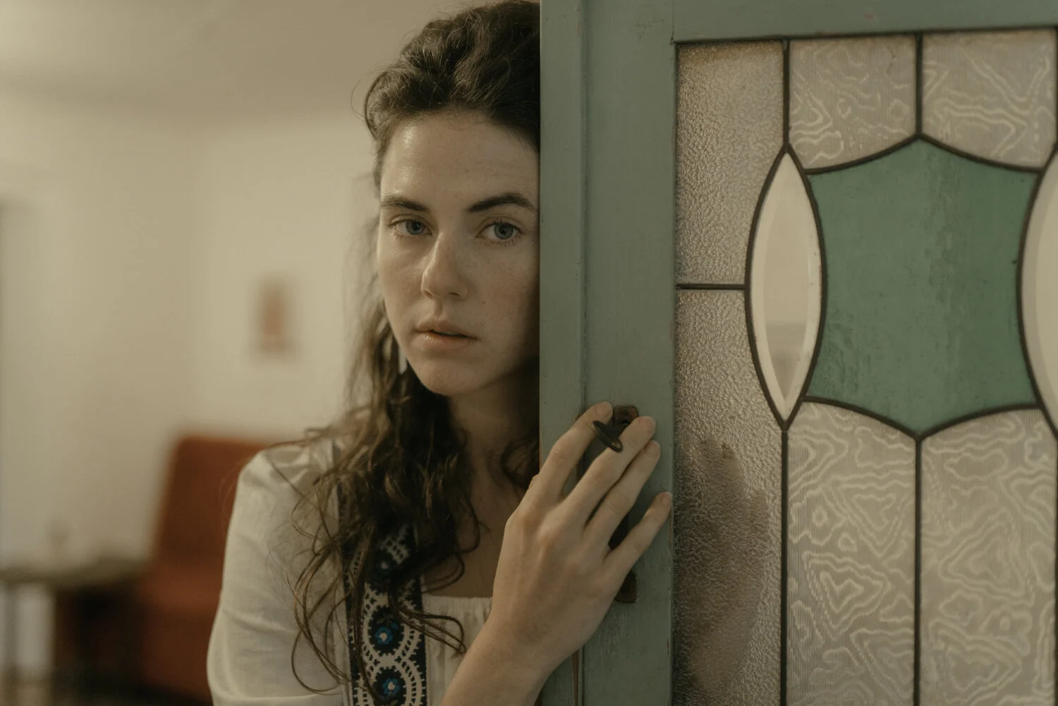 Young woman with long dark hair peeking out from behind a door, holding the door handle with her hand, in a room with a red chair in the background.