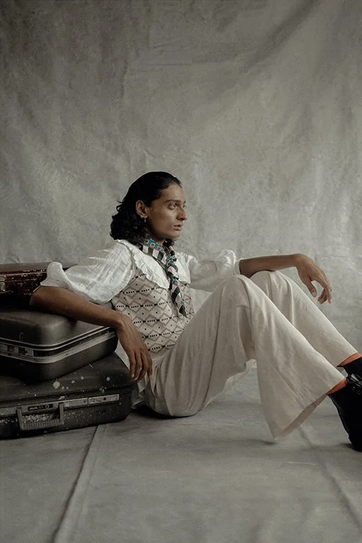 A woman with dark, curly hair sitting on the floor, leaning against a suitcase, with an expressionless face. She is wearing a white blouse, beige pants, and black shoes, in front of a plain, textured wall.