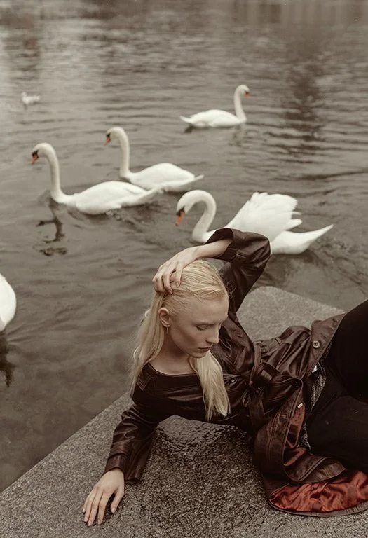 A young woman with long blond hair reclining on a concrete ledge near a body of water with seven swans swimming behind her.