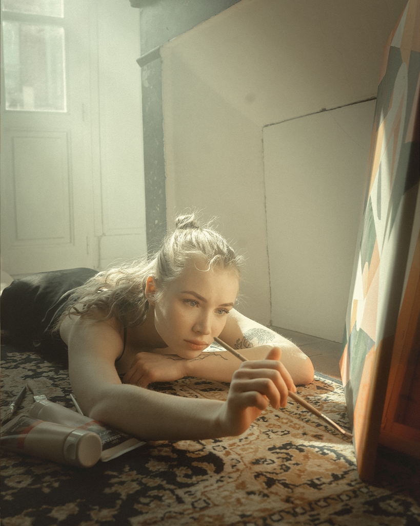 Woman lying on floor, painting on canvas in a sunlit room.