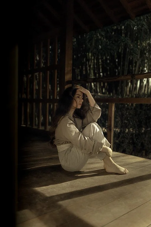 A woman sits on a wooden floor inside a rustic wooden cabin, with her hand resting on her forehead and her knees pulled up. Sunlight streams through the open window, illuminating the room and creating shadows on the floor. Outside, trees are visible 