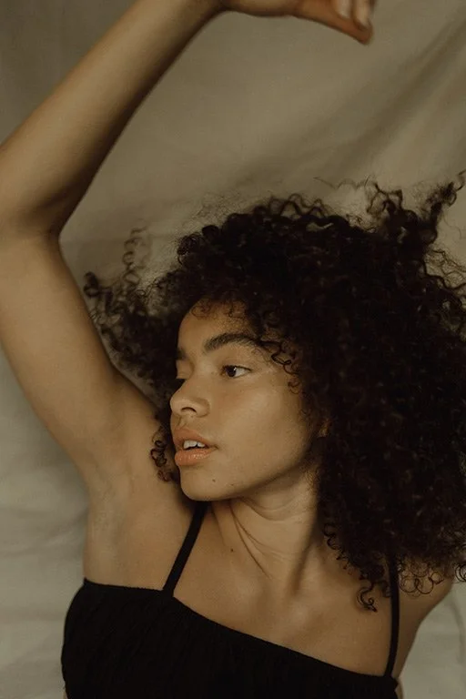 A woman with curly hair lying on her side with one arm raised above her head, wearing a black spaghetti strap top.