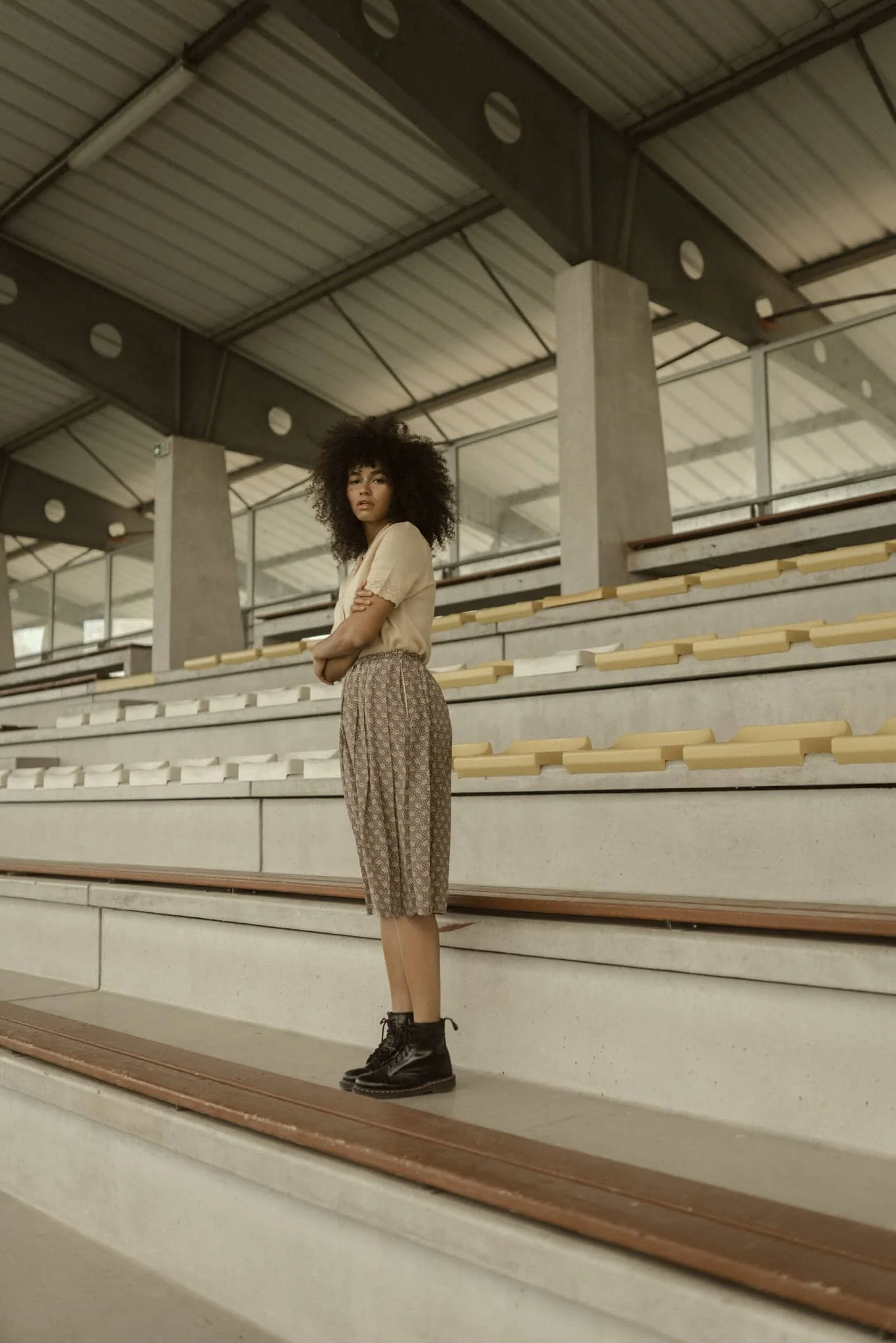 Young woman with curly hair standing on stadium bleachers indoors, wearing a beige shirt, patterned knee-length pants, and black boots.