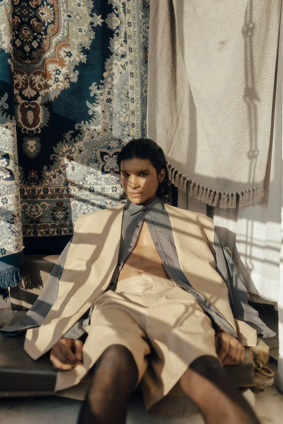 A young man sitting on a bench in front of patterned curtains and white textured drapes, wearing beige shorts, sheer black tights, an open beige blazer, and a striped shirt with the collar wide open. Sunlight casts shadow patterns across his face and