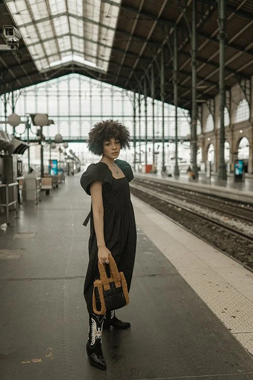 A woman standing on a train platform in a train station, holding a woven bag, wearing a black dress and cowboy boots.