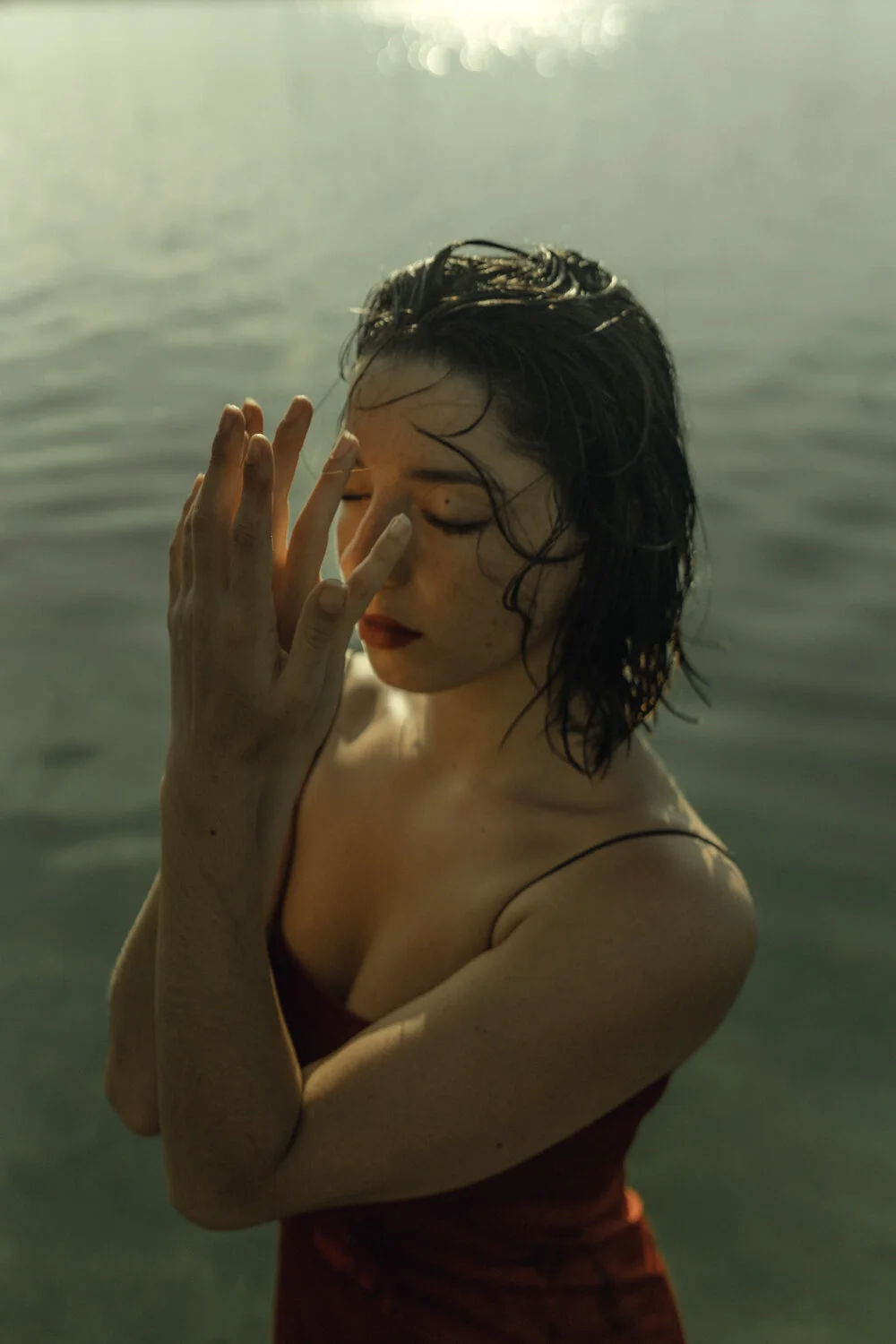 A woman with wet hair and closed eyes stands in water, gently touching her face with her fingers.