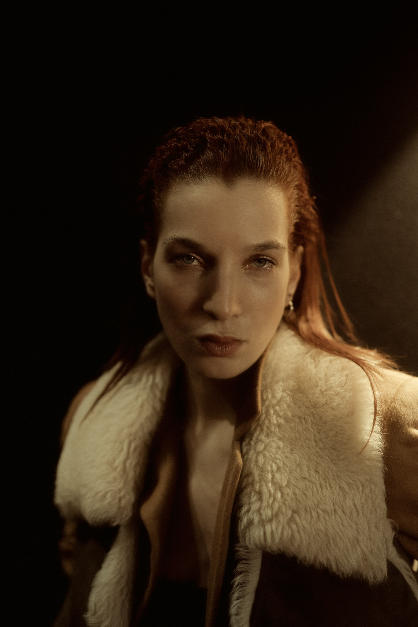 Portrait of a woman with red hair wearing a beige shearling jacket against a dark background.