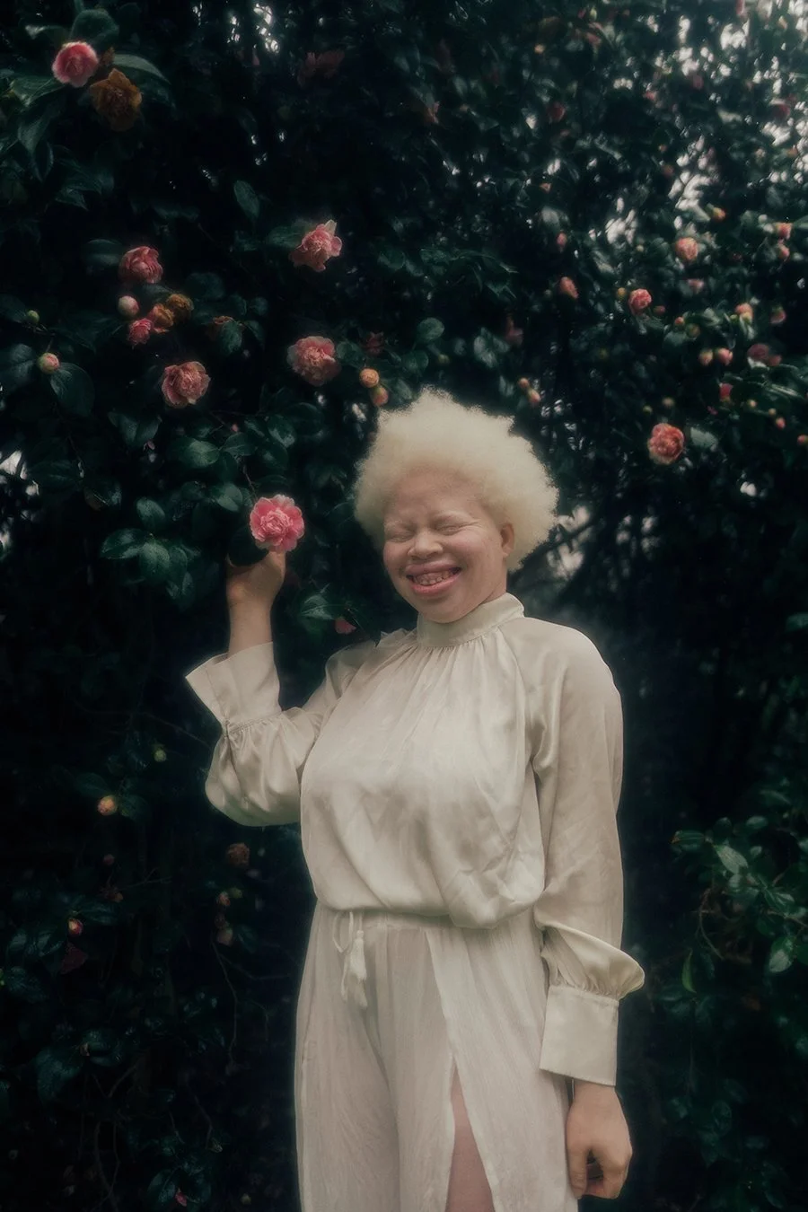 A person with white curly hair smiling and sticking out their tongue, wearing a cream-colored outfit, standing among dark green shrubbery with pink flowers.