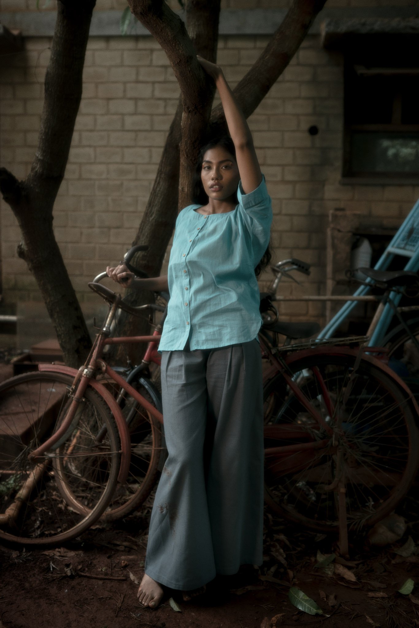 A woman in a light blue blouse and wide gray pants standing near a tree with old bicycles in the background.