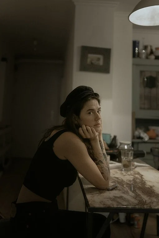 A young woman with dark hair, wearing a black top and a black beret, sitting at a wooden table with her chin resting on her hand, looking at the camera in a dimly lit kitchen.