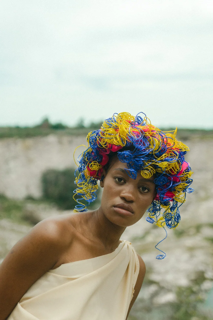 A woman with a colorful, curly headwrap made of yellow, blue, and pink yarn, wearing a beige off-shoulder dress, standing outdoors in a natural landscape.
