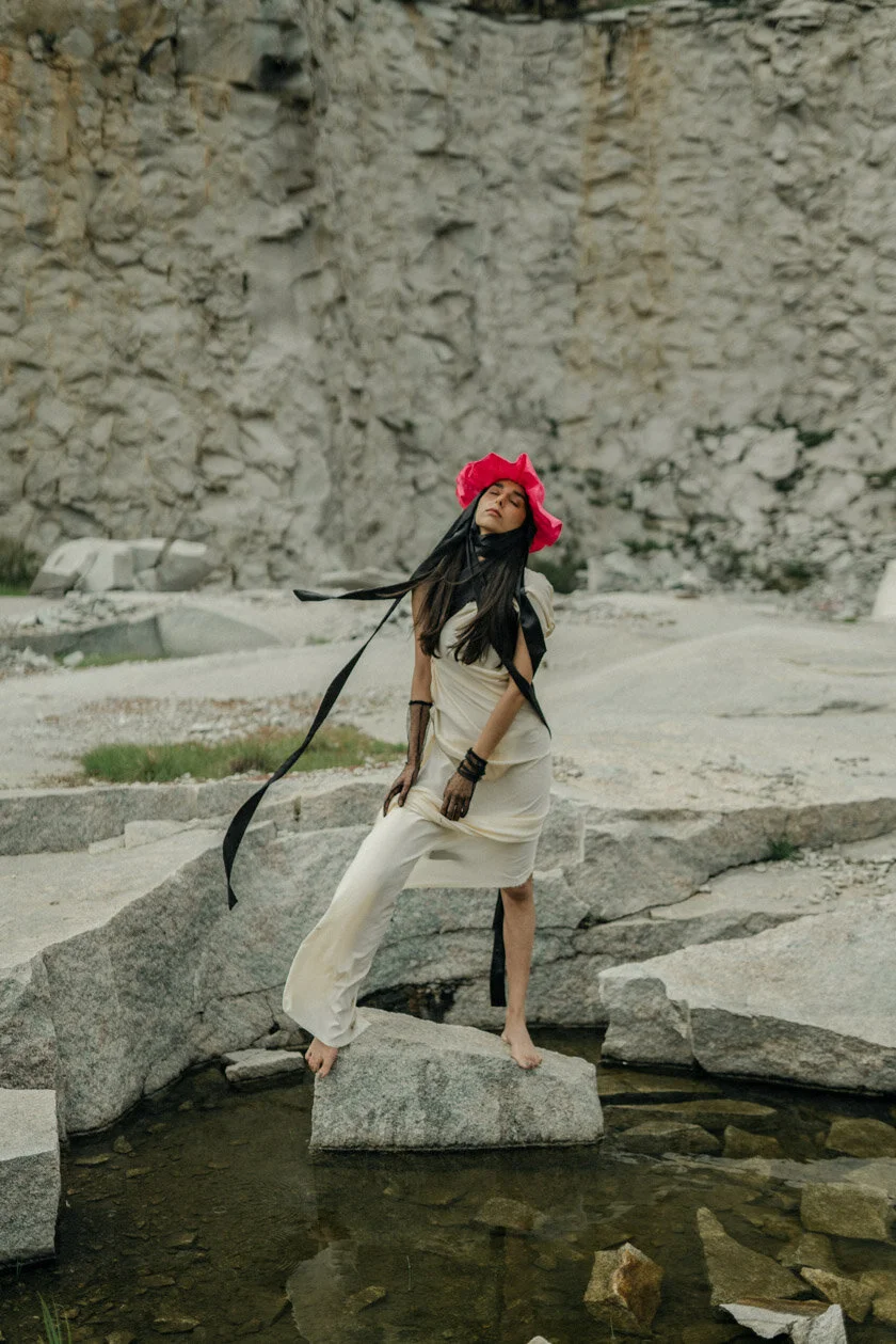 A woman in a light-colored dress and black gloves poses barefoot on rocks by a small body of water outdoors, wearing a bright pink hat and a black scarf flowing in the wind against a rocky background.